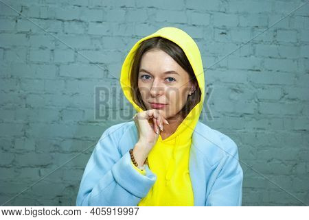 Woman Thinking With Hand Over Chin And In Yellow Hood On Head Standing Isolated Over Blue Brick Back