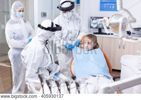 Little Girl Looking At Dentist While Assistant Is Putting Her A Bib Before Dental Examination Uring 