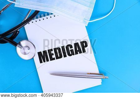 Medigap, Text Written In A Notebook Lying On A Blue Background, With A Stethoscope And A Medical Mas