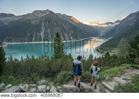 Olpererhutte, Austria - Aug 7, 2020: Two Female Trekkers Take Photo Of Glacier Reservoir At Sunset H