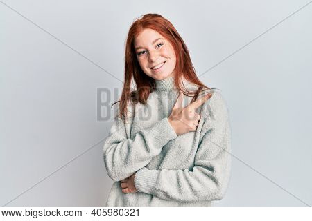 Young red head girl wearing casual winter sweater smiling cheerful pointing with hand and finger up to the side 