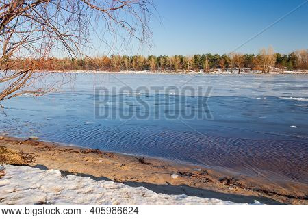 Section Of Wide Plain River Partially Covered With Thin Ice, Willow Branches On A Foreground Against