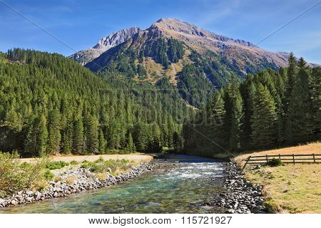  Austrian Alps. Headwaters Krimml waterfalls. The narrow stream flows between fields and pine forests. Bluish - green transparent water glows in the sun