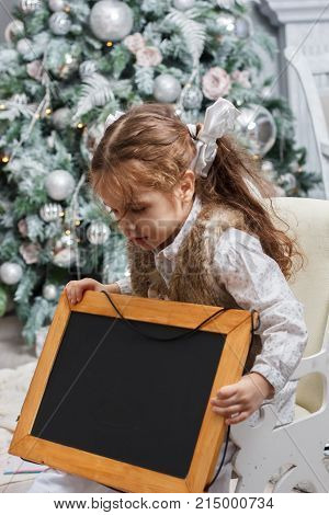 Little kid looks to black board in shees hands. Christmas tree as background