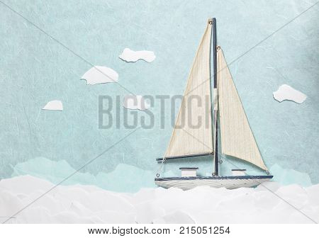 Wooden Sailboat Model on light blue background with sea made from paper. Yacht model with clouds made from papers shoot from above