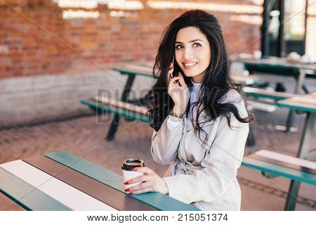 Gorgeous dark-haired woman with pure skin, dark bright eyes and full lips holding takeaway coffee and smartphone chatting with her boyfriend having pleased look. Young female enterpreneur resting