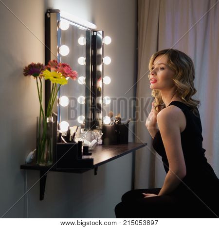 Attractive Blonde Girl Primp At The Mirror With Lamps In The Beauty Studio. A Happy Satisfied Custom