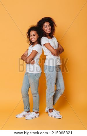Full length portrait of two happy african sisters standing back to back with arms folded isolated over orange background