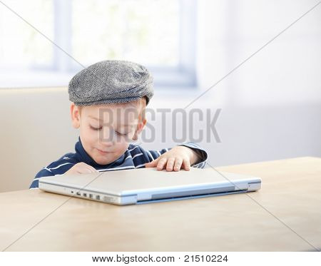 ¿Dulce niño pequeño sentado en la mesa, con tapa, jugando con el ordenador portátil.?