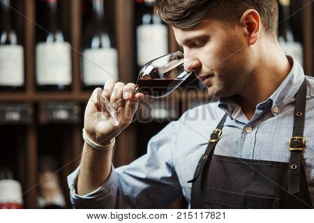 Sommelier smelling flavor of red wine in bokal on background of shelves with bottles in cellar. Male appreciating color, quality and sediments of drink. Professional degustation expert in winemaking.
