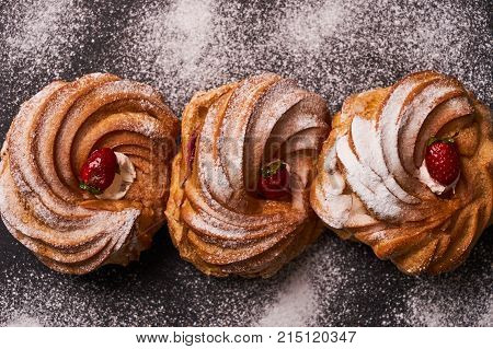 Profiterole, french puff pastry eclairs with custard cream, strawberry and powdered sugar on dark slate cake board on blue background, close-up, top view.
