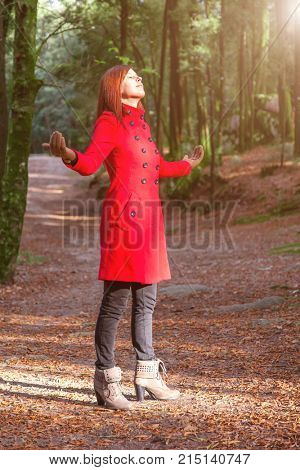 Woman enjoying the warmth of winter sunlight alone on forest park path with arms open receiving rays of light, wearing a red long coat or overcoat