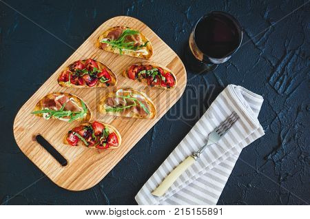 Brushetta set for wine. Variety of small sandwiches with prosciutto tomatoes arugula fresh basil and balsamic sauce served on wooden board with glass of red wine over dark background. Top view.