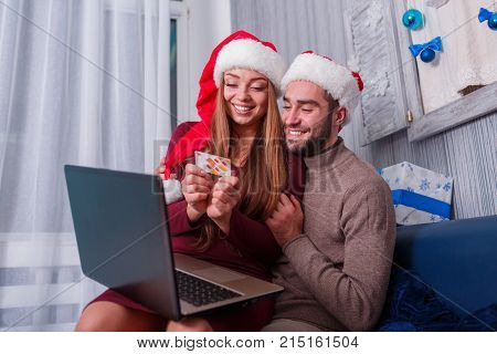 Happy young couple doing online shopping, on New Year's Eve, sitting on sofa at home, using credit card and laptop.