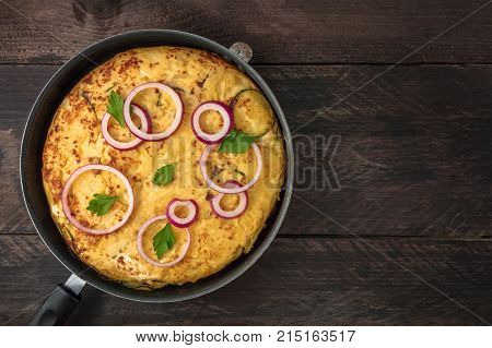 An overhead photo of a Spanish tortilla with potatoes and zucchinis in a tortillera, on a dark rustic wooden background with a place for text
