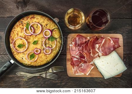An overhead photo of a Spanish tortilla with potatoes and zucchinis in a tortillera, with jamon, cheese, glasses of white and red wine