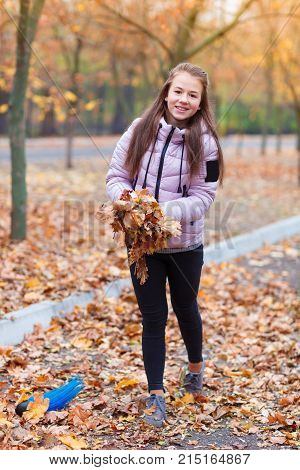 A girl in a pink jacket, standing in an autumn park, holds a bunch of leaves in her hands, collects the fallen leaves.