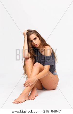 A young, sexy girl sits with bent knees in a gray T-shirt and shorts on a white background