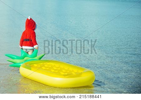 Christmas happy child with pineapple mattress in water. Winter holiday vacation. New year small boy at beach. Xmas party celebration childhood. Santa claus kid in red Christmas costume.