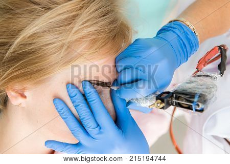 Cosmetologist applying permanent make up on eyebrows. Beautiful woman with smooth young skin sitting in a beautician. A beautician performs modeling permanent eyebrows makings.