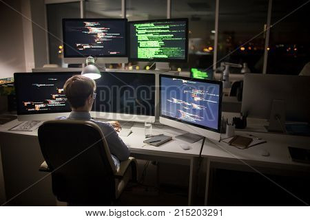 Back view of highly professional coder looking at wristwatch while sitting in front of computer and working on ambitious project, interior of dim open plan office on background
