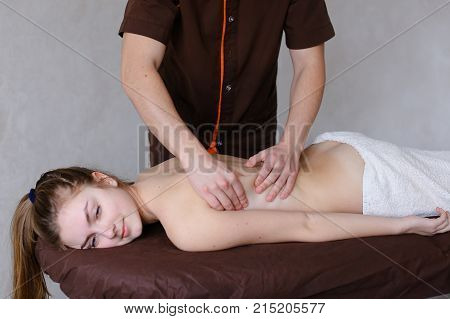 Friendly young man masseur concentrated and gently makes useful medical procedure and drives hands along back to nice girl who came to reception and lies on couch to half covered by terry towel in light cosmetology salon. Man of European appearance with s