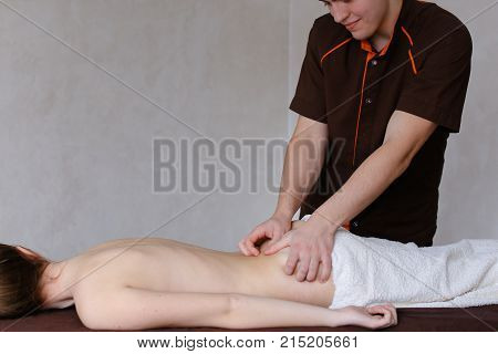 Friendly young man masseur concentrated and gently makes useful medical procedure and drives hands along back to nice girl who came to reception and lies on couch to half covered by terry towel in light cosmetology salon. Man of European appearance with s
