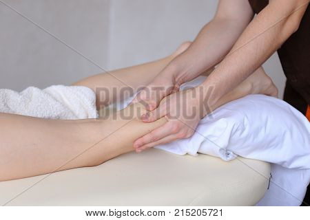Clever guy massage therapist gently massages feet and rubs palms for effect of relaxation, which came to reception of young woman who lies on couch covered with terry towel in light cosmetology salon. Experienced male doctor of European appearance with sh