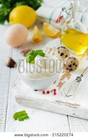 Traditional mayonnaise sauce in white ceramic bowl and ingredients for its preparation on white wooden background. Selective focus.