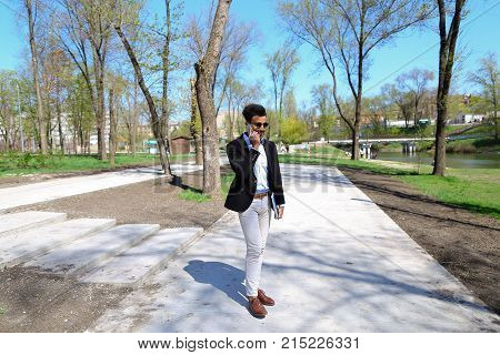 Guy walking in park and smiling, slow motion. Man dressed in blue shirt, white jeans and brown shoes. Boy has black hair and beard. Concept of places for rest and clean parks and cheeps fashion clothes for man