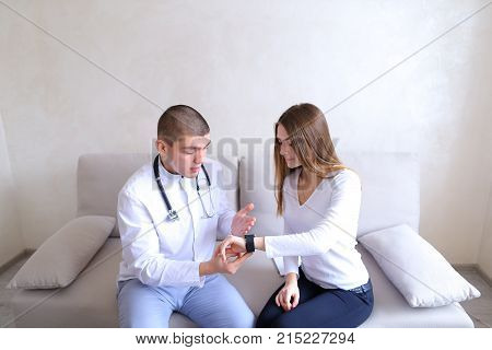 Successful male cardiologist who shows female on iWatch gadget, which girl on arm has to control analysis and adjustment of day's routine. Therapist with stethoscope and patient sitting on couch on background of gray wall in hospital's office during day. 