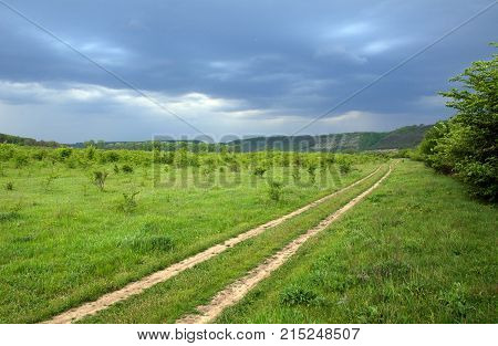 Road Along The Green Valley With Bushes And Trees. Rock On The Horrizon, Blue And Grey Stormy Clouds