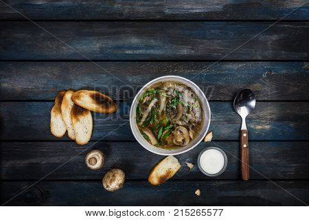 Fresh mushroom soup with green onion sour cream and bread crisps. In white bowl dishe. Colored wooden background, spoon. Top view.