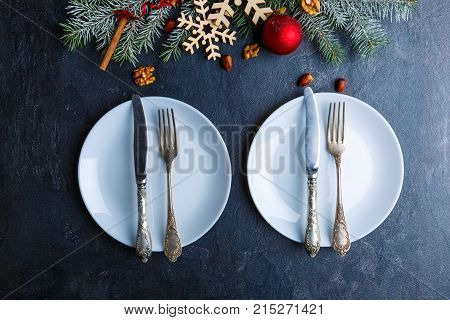 On the dark surface there are two white round plates with knives and forks, next there is a decoration made of a Christmas tree branch with wooden snowflakes, Christmas tree toys and nuts. View from above. Indoors.