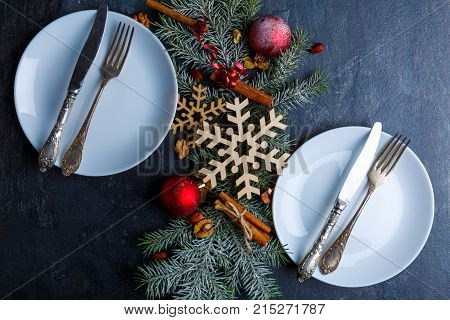 Christmas tree branch decorated with wooden snowflakes, Christmas balls and cinnamon sticks is between two white round plates with cutlery. Close-up. View from above. Indoors.