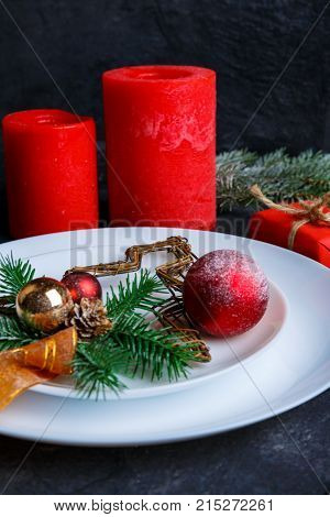 In a white round plate there is a decorated Christmas tree with balls and ribbon, next to two large red candles. Christmas decor. Indoors.