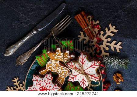 Next to a plate with sweet Christmas gingerbread cookies of different shapes in multi-colored sugar glaze cutlery and decorations from a Christmas tree branch, wooden snowflakes and cinnamon sticks. View from above.