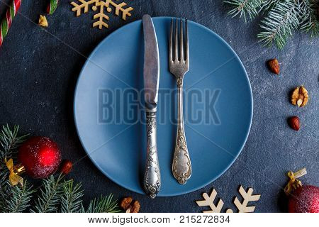 On a blue round plate, an old knife and fork, around Christmas decorations from Christmas tree branches, wooden snowflakes and red balls.