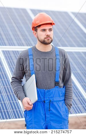 A serious worker at the solar battery station with a beard, dressed in a blue uniform and an orange helmet, keeps a modern tablet on hand and looks somewhere with a serious look. Outdoors.