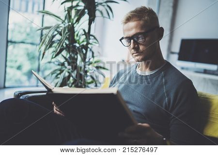 Portrait handsome young man wearing glasses casual clothes holding book hands.Man sitting in vintage armchair modern loft studio reading book. Blurred background.Horizontal