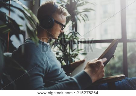 Attractive young man wearing glasses casual clothes.Man sitting in vintage armchair modern loft studio, reading book and relaxing whith headphone music. Blurred background.Horizontal