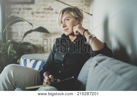 Beautiful young woman writing something in note pad while sitting on armchair at living room.Charming female studying doing homework at home.Horizontal.Blurred background