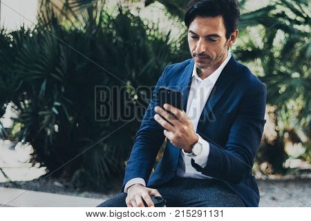 Attractive smiling businessman making video conversation with partners by mobile phone.Man using contemporary smartphone sitting at city park.Blurred background. Horizontal