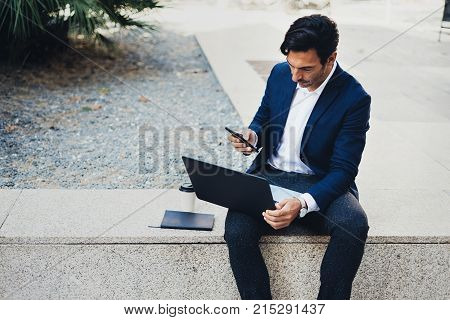 Attractive young businessman using contemporary notebook and smartphone sitting outside.Blurred background. Horizontal