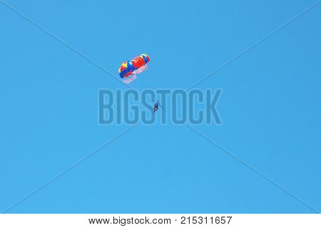 Two people are flying on a large color parachute. A bright sunny day. Blue glowing sky. Parachute red blue yellow and white. The color of the parachute contrasts strongly with the sky.