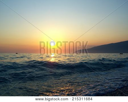 Bright bright sun. Calm sea. Not large waves roll on a gentle beach and roll back into the sea. Over the sea after a hot day hangs a thick haze. The rays of the sun in the haze turn red. In the distance a mountainous shore overgrown with forest is visible
