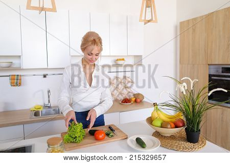 Attractive young woman cuts green cucumber for salad with sharp knife, takes red juicy tomato and several times throws up with smile on face, sniffs freshness of lettuce leaves and takes up shredding vegetables. Blonde at top of modern light kitchen with 