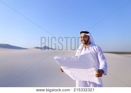 Beautiful Emirate Sheikh businessman studying project of future construction of complex, standing in middle of bottomless desert with white clean sand on clear warm day against blue sky. Swarthy Muslim with short dark hair dressed in kandura, long, spacio