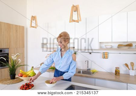 Slender, sweet woman cooks delicious breakfast of oatmeal and ripe red strawberry, with smile on face, takes plate in hands and brings it to camera lens, standing in stylish and light kitchen with white cabinets, household appliances and variety of utensi