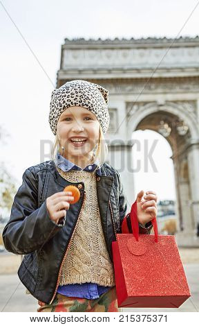 Stylish autumn in Paris. Portrait of smiling elegant child with red present bag near Arc de Triomphe in Paris holding French macaroon